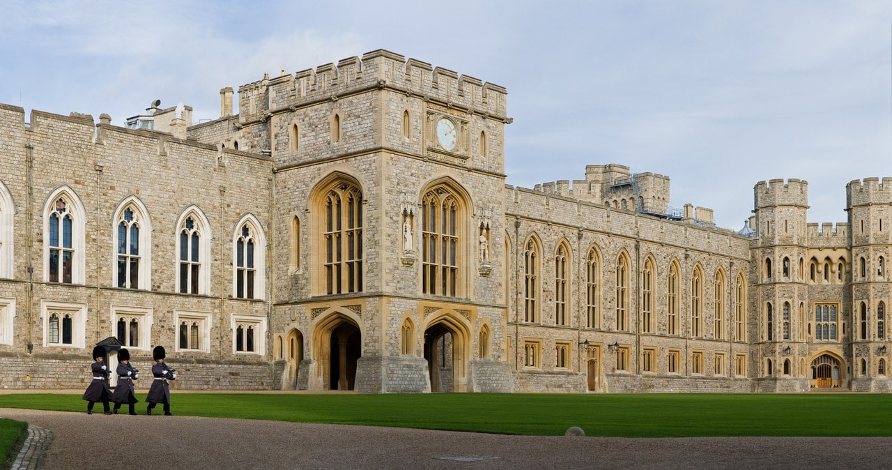 A long battlemented range round the Upper Ward quadrangle, a clock tower above an arcade of pointed arches, with three guardsmen in bearskins on the grass A long battlemented range round the Upper Ward quadrangle, a clock tower above an arcade of pointed arches, with three guardsmen in bearskins on the grass