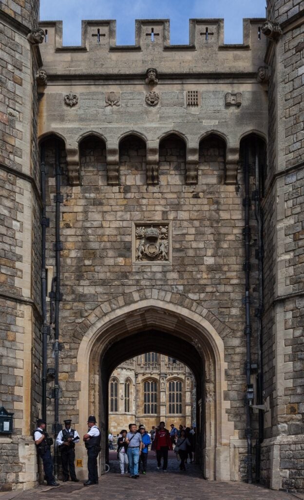 Windsor Castle 6 The outer face of a machicolated stone gateway, carved badges and a royal coat of arms above the pointed arch, with the traceried windows of St George's Chapel visible through it