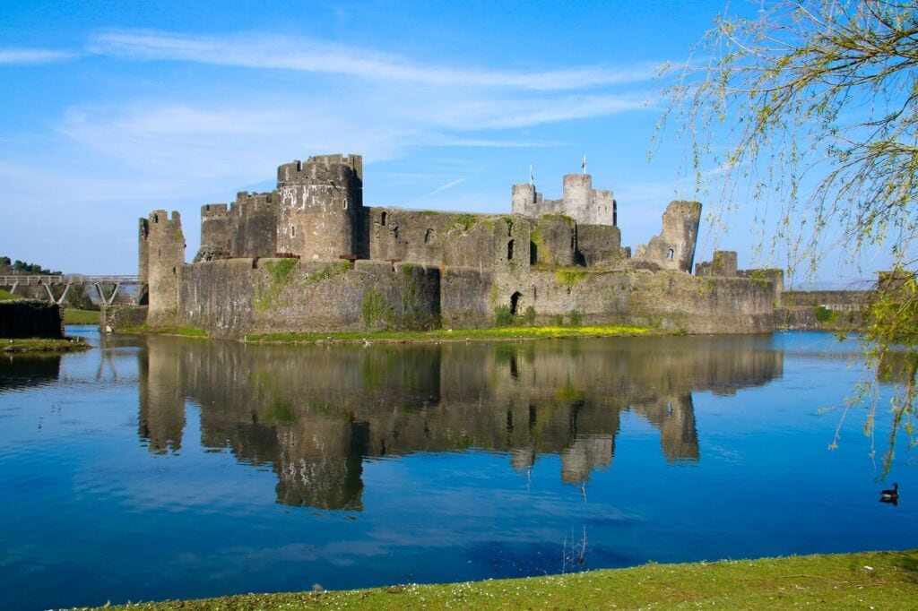 Caerphilly Castle 5 Caerphilly Castle's towers and walls reflected in a calm lake under a blue sky, with willow branches at the right edge