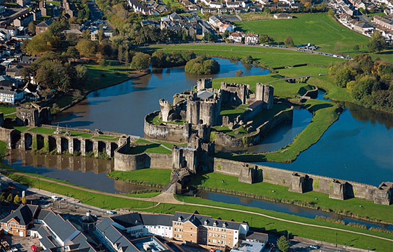 Aerial view of Caerphilly Castle with a long fortified wall and gatehouse across the foreground, an arcaded section holding back water at the left, and the castle on an island in broad lakes beyond Aerial view of Caerphilly Castle with a long fortified wall and gatehouse across the foreground, an arcaded section holding back water at the left, and the castle on an island in broad lakes beyond
