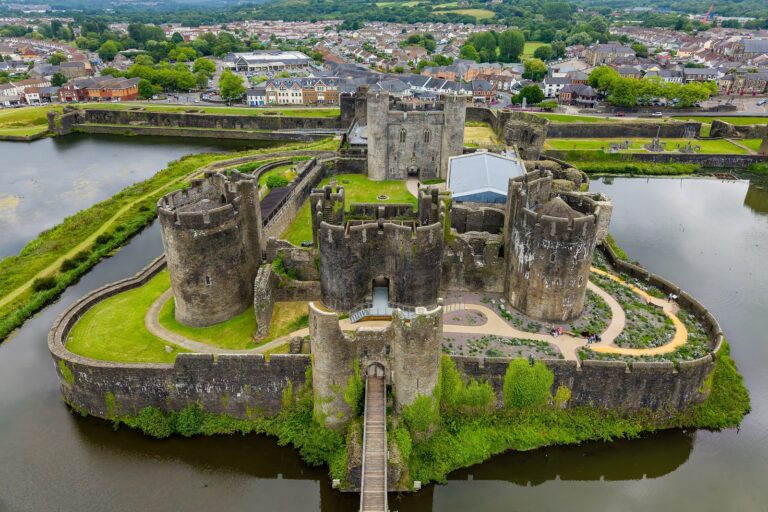 Drone view of Caerphilly Castle on its island: a high inner ward with round corner towers and two twin-towered gatehouses, inside a lower curtain wall, with water on every side and the town beyond