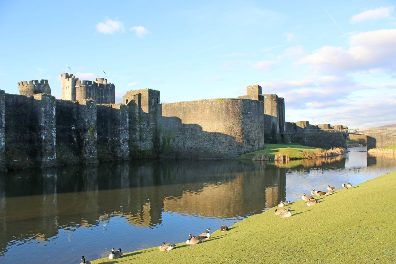 A long stone wall stiffened by a row of tall square buttresses, rising from still water that reflects it, with geese on the grassy bank in the foreground A long stone wall stiffened by a row of tall square buttresses, rising from still water that reflects it, with geese on the grassy bank in the foreground
