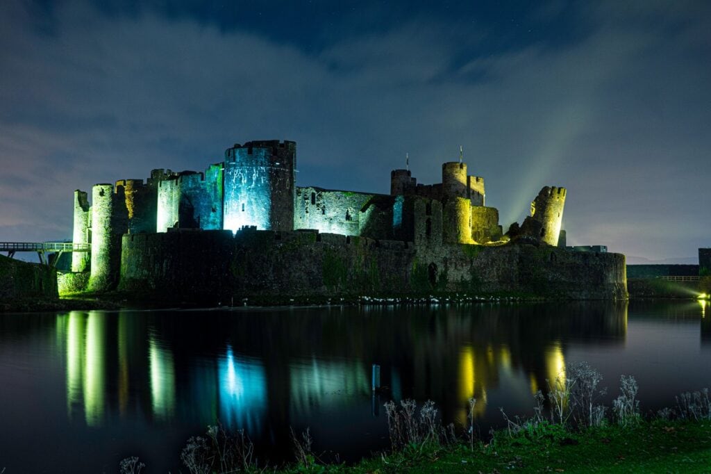 Caerphilly Castle 7 Caerphilly Castle at night, its towers lit in colored light and reflected in black water under a dark blue sky