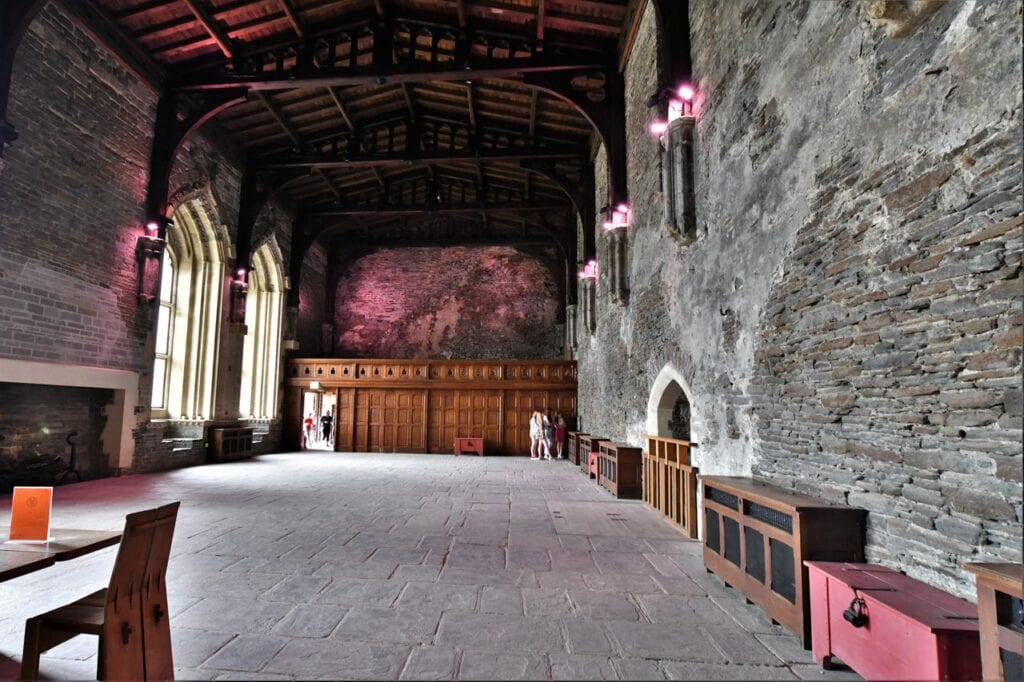 Caerphilly Castle 6 A long, high hall with a dark arched timber roof, tall pointed windows down the left side, a stone-flagged floor, a timber screen with a doorway at the far end, and wooden chests and benches along the right-hand wall