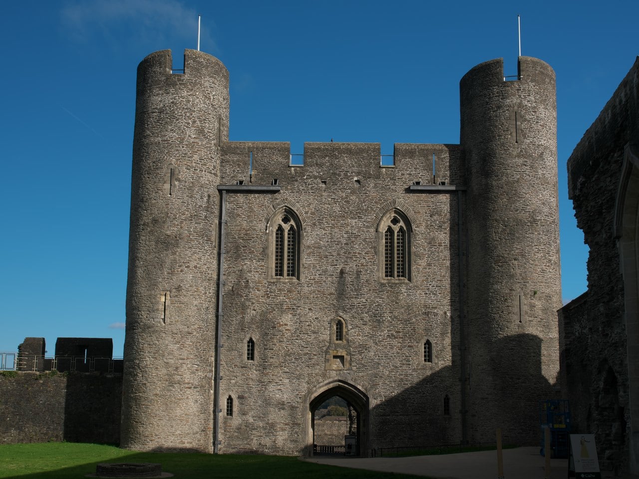 A tall stone gatehouse with a tall round turret at each corner of its courtyard face, two pointed traceried windows in the center, and below them a small window above a stone-framed opening over a pointed arch at ground level, against a deep blue sky A tall stone gatehouse with a tall round turret at each corner of its courtyard face, two pointed traceried windows in the center, and below them a small window above a stone-framed opening over a pointed arch at ground level, against a deep blue sky
