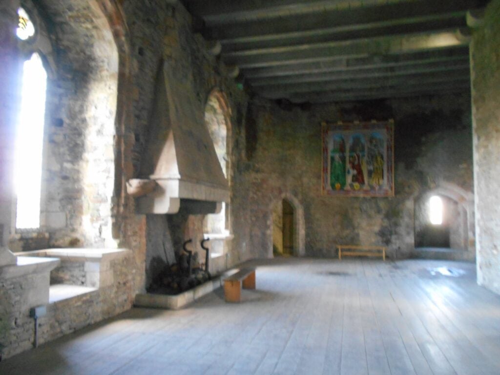 Caerphilly Castle 8 A long stone room with a timber-beamed ceiling and plank floor, a large hooded stone fireplace at the left, deep window embrasures and a colorful painted hanging on the end wall