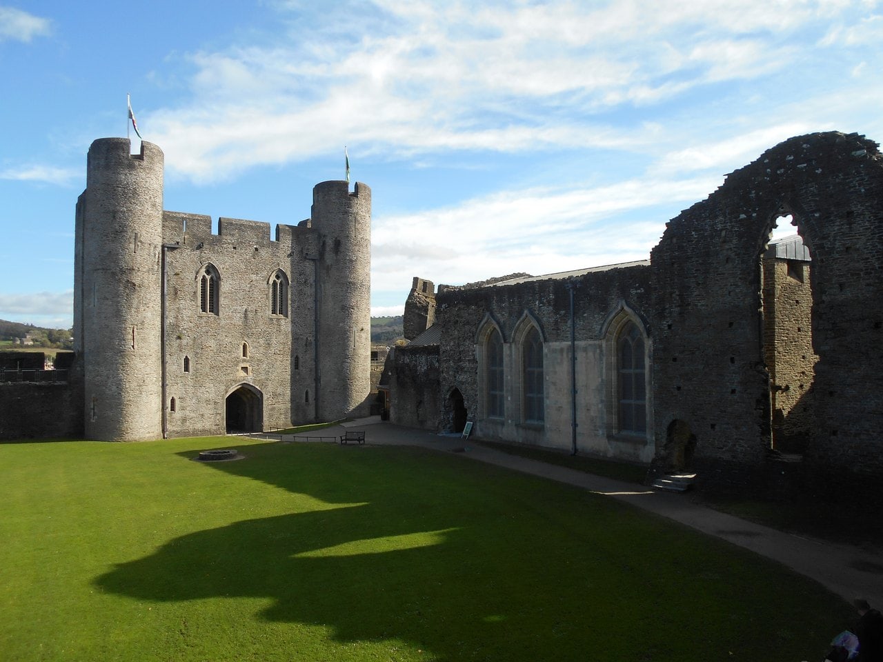 A grassy courtyard enclosed by stone walls, a twin-towered gatehouse at the far end and a long hall range with tall pointed windows and a ruined gable at the right A grassy courtyard enclosed by stone walls, a twin-towered gatehouse at the far end and a long hall range with tall pointed windows and a ruined gable at the right