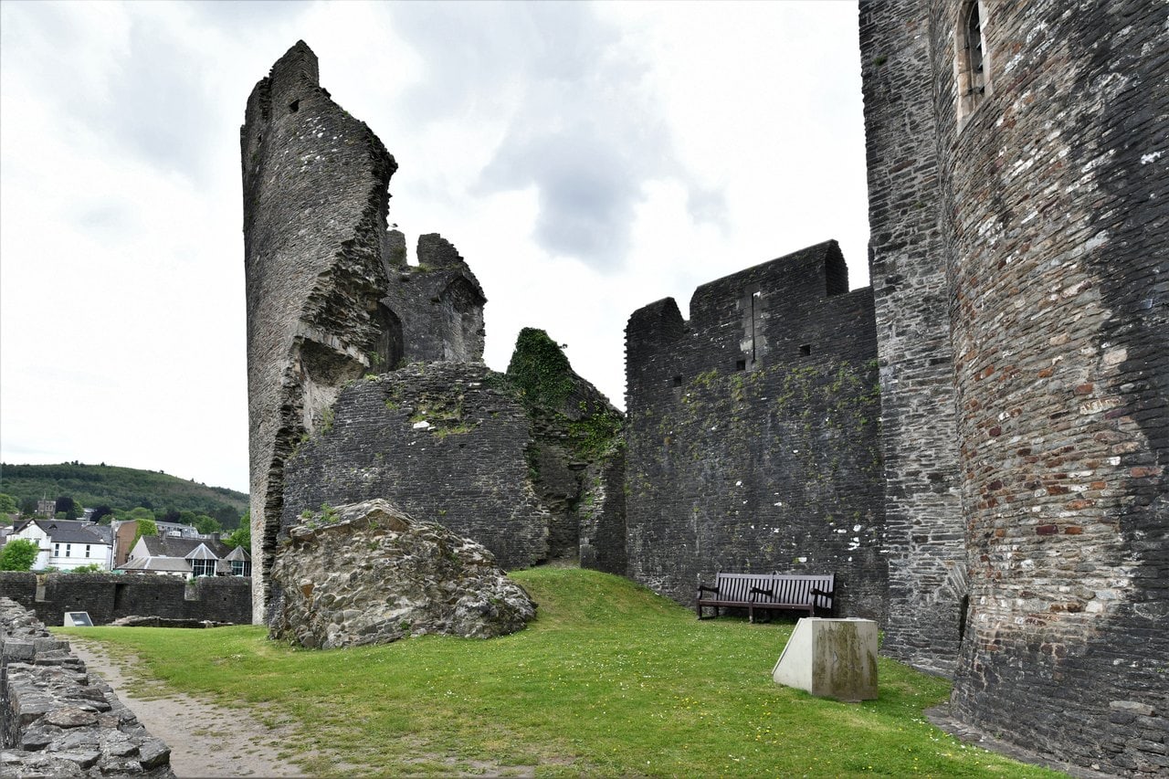 A broken round stone tower tilting visibly out of vertical beside lower walls and a fallen block of masonry, on a lawn under a gray sky A broken round stone tower tilting visibly out of vertical beside lower walls and a fallen block of masonry, on a lawn under a gray sky