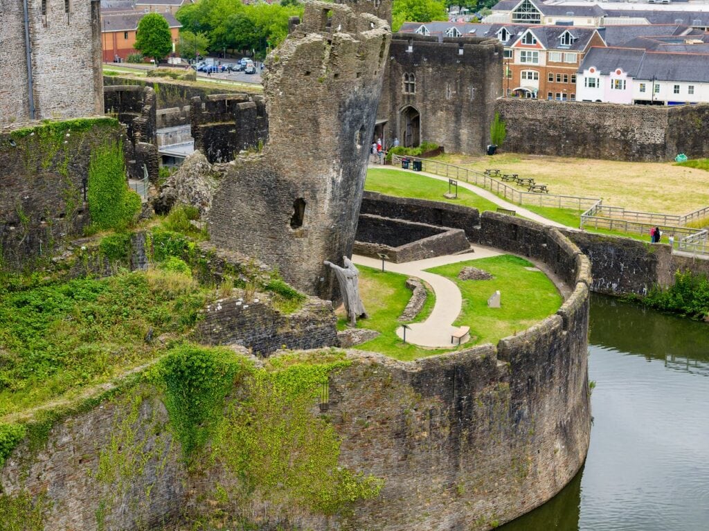 Caerphilly Castle 3 Aerial view of a broken round tower tilting outward behind a curved wall above the water, a pale standing sculpture at its base and a lawn with a looping path beside it