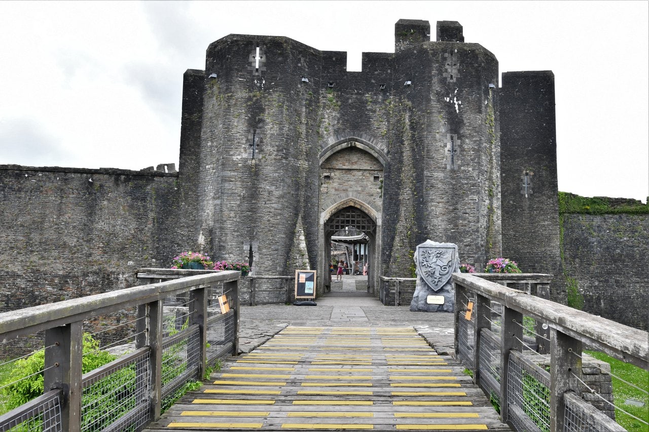 A massive stone gatehouse with two many-sided towers and a pointed entrance arch, reached by a timber footbridge with handrails, flower baskets at the gate A massive stone gatehouse with two many-sided towers and a pointed entrance arch, reached by a timber footbridge with handrails, flower baskets at the gate