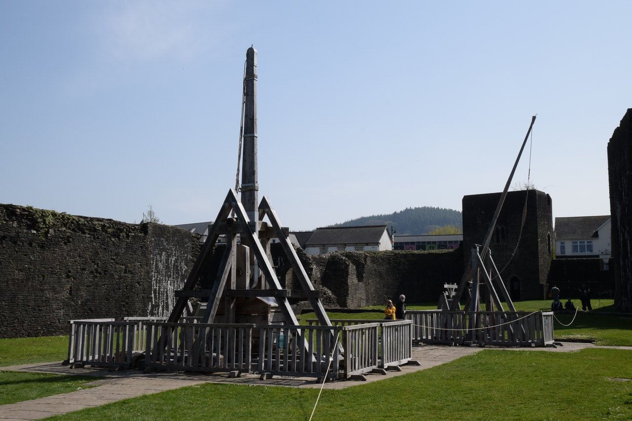 Two tall timber siege-engine replicas inside wooden fences on a lawn, with a curtain wall and a square tower behind Two tall timber siege-engine replicas inside wooden fences on a lawn, with a curtain wall and a square tower behind