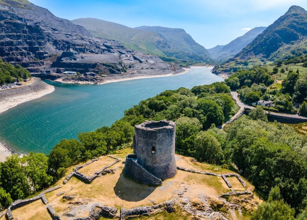 Dolbadarn Castle 4 Drone view of a round stone tower on a sunlit knoll above a turquoise lake, with terraced gray slate quarries on the far shore and a mountain valley beyond
