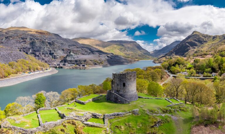 Drone view of Dolbadarn Castle: a round stone tower and low wall footings on a green knoll, with a lake, gray slate-quarry terraces and mountains behind