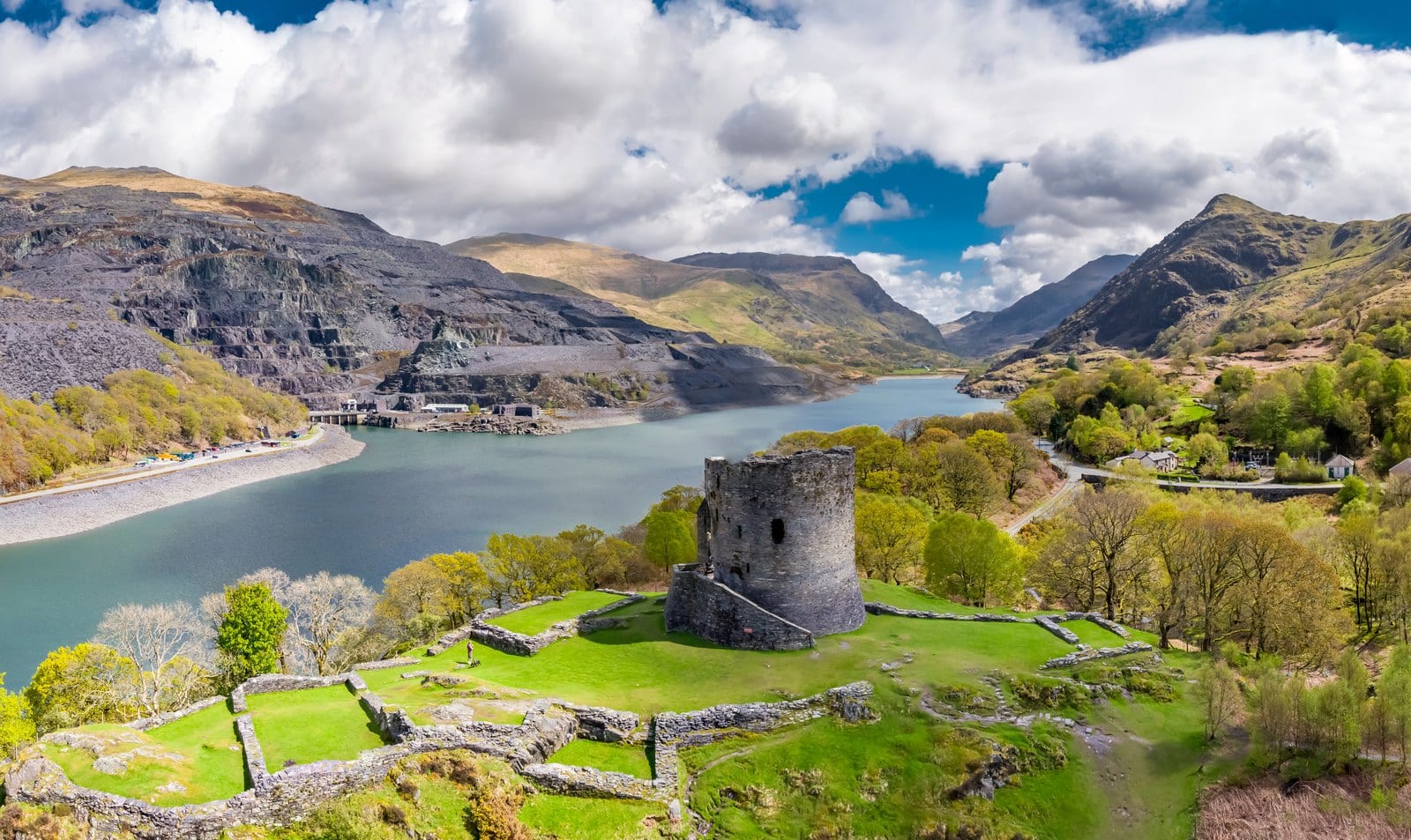 Dolbadarn Castle 1 Drone view of Dolbadarn Castle: a round stone tower and low wall footings on a green knoll, with a lake, gray slate-quarry terraces and mountains behind