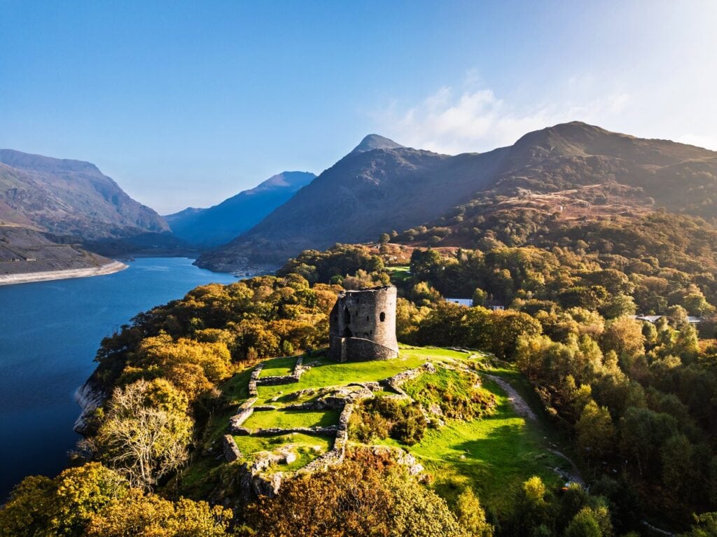 Dolbadarn Castle 2 Drone view in autumn of a round stone tower and low wall footings on a knoll among orange and green trees, with a lake to the left and mountains beyond