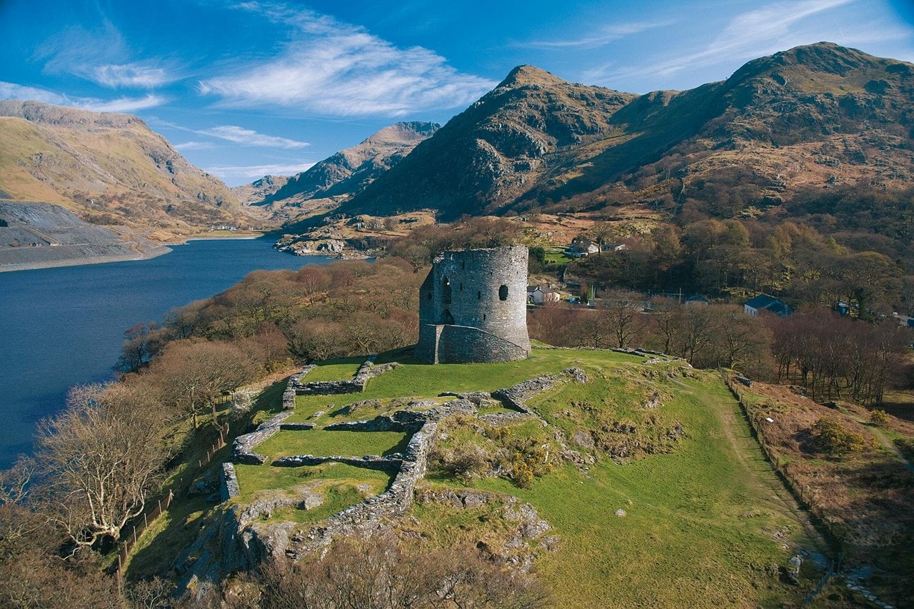 Aerial view of a round stone tower on a green knoll with the low outlines of stone buildings along its edge, a lake on the left and mountains under blue sky Aerial view of a round stone tower on a green knoll with the low outlines of stone buildings along its edge, a lake on the left and mountains under blue sky