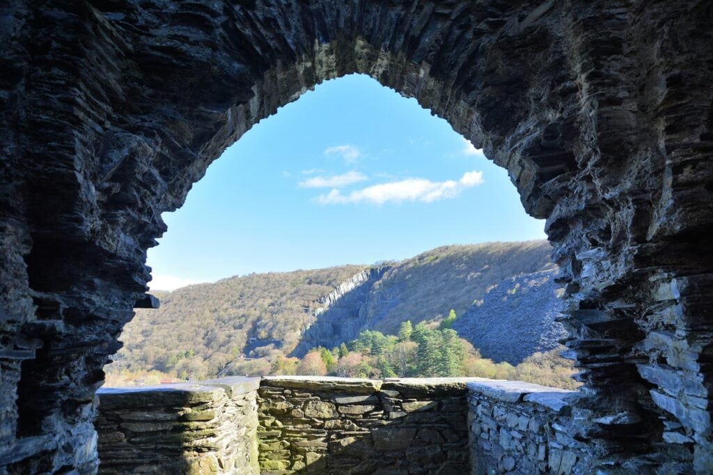 Dolbadarn Castle 6 Looking out through a pointed stone arch onto a small walled stone landing, with a wooded hillside and gray quarry spoil beyond under blue sky