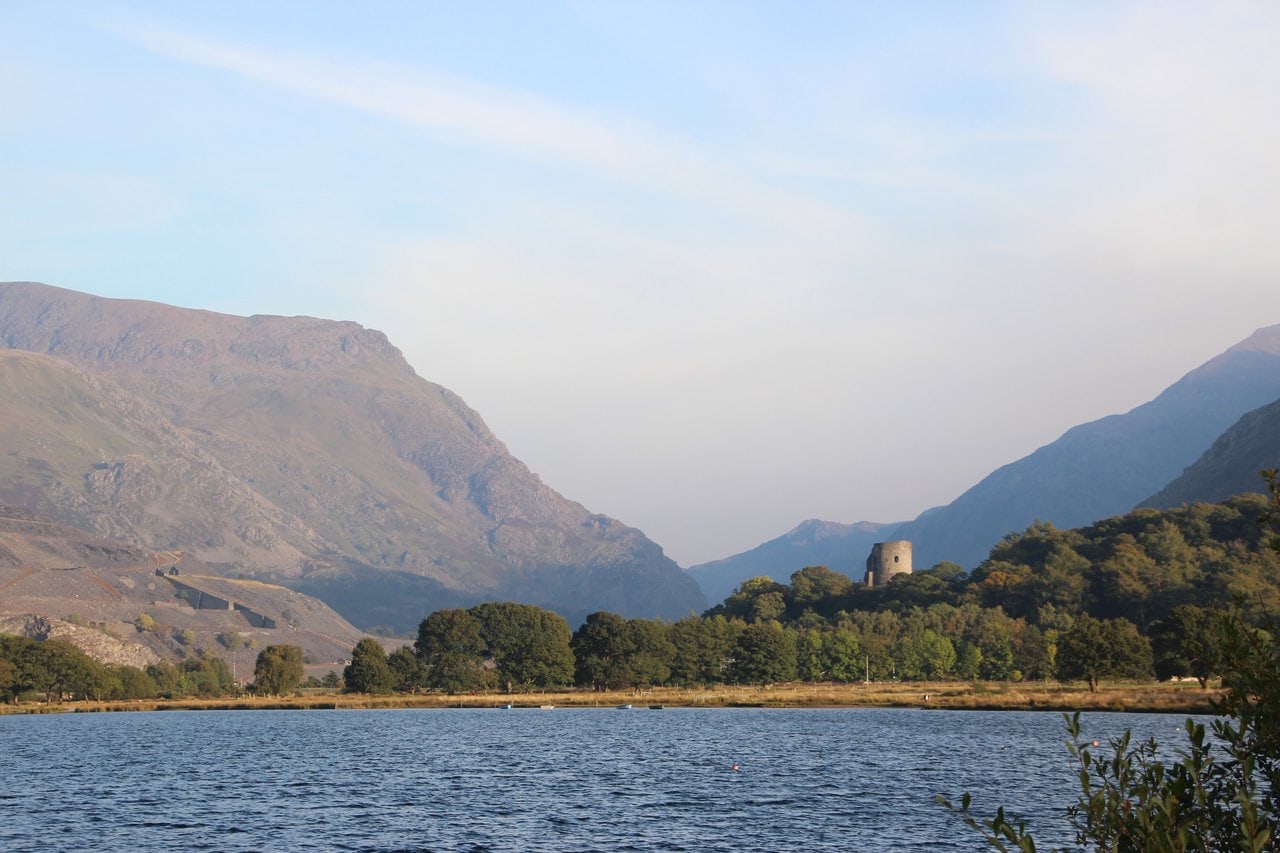A distant round stone tower on a wooded knoll beyond a lake shore, set between two steep mountainsides that open into a long valley under a hazy sky A distant round stone tower on a wooded knoll beyond a lake shore, set between two steep mountainsides that open into a long valley under a hazy sky