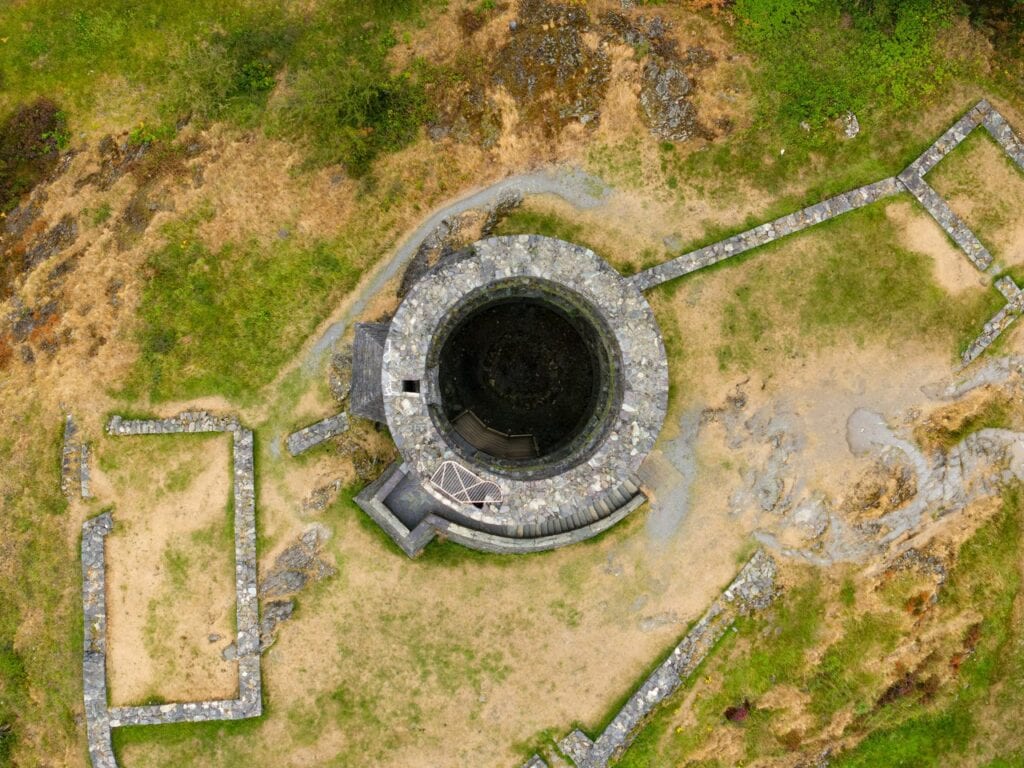 Dolbadarn Castle 3 Straight-down drone view of a hollow round stone tower with a curved block of masonry against one side, surrounded by grass and the low outlines of rectangular stone foundations