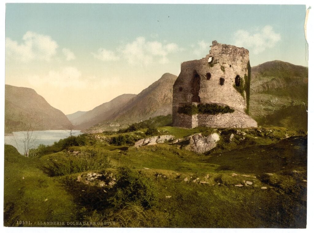 Dolbadarn Castle 15 Hand-colored late-nineteenth-century photochrom of a ruined round tower with ivy on its walls, on a grassy rocky knoll, with a lake and mountains behind