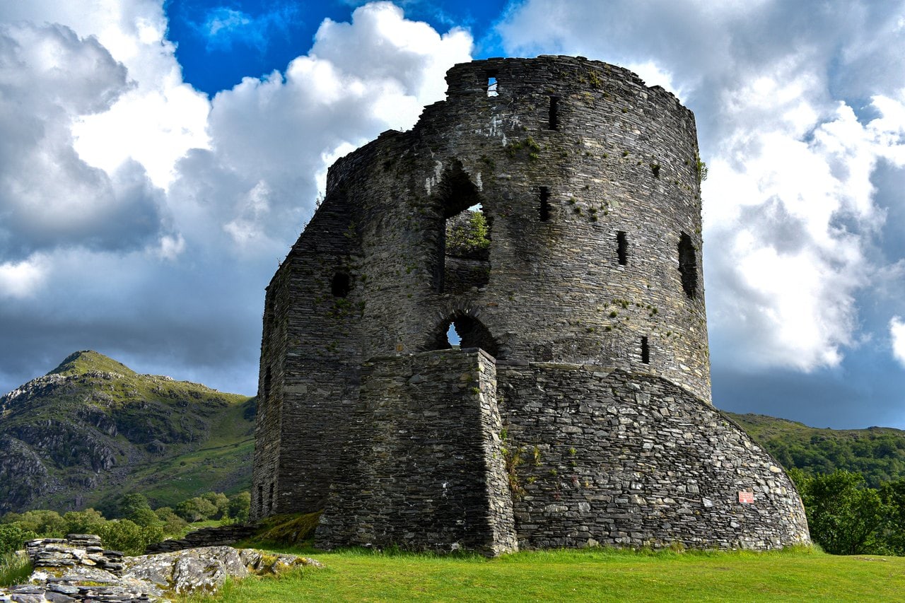 A tall round tower of dark stone rising from a curved masonry base on green grass, with a pointed doorway, a second, larger pointed opening with broken sides above it and narrow slit windows, against blue sky and white cloud A tall round tower of dark stone rising from a curved masonry base on green grass, with a pointed doorway, a second, larger pointed opening with broken sides above it and narrow slit windows, against blue sky and white cloud