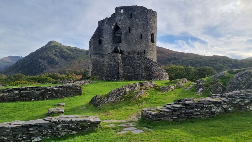 Dolbadarn Castle 12 Low walls of flat stacked slate stones on green grass in the foreground, with the ruined round tower and a mountain behind under a cloudy sky