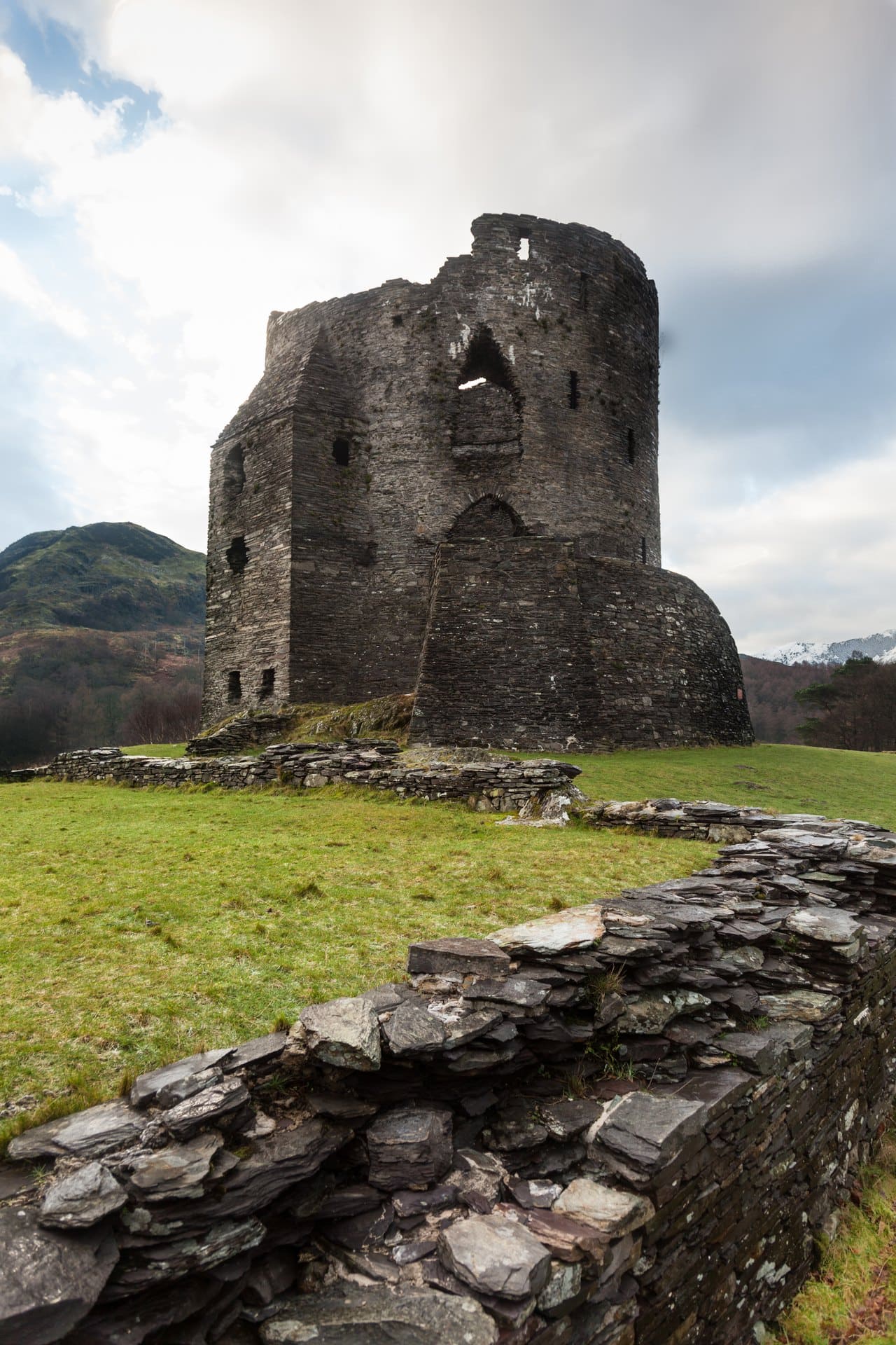 Portrait view of a low wall of flat, dark slate slabs running diagonally in the foreground across grass toward a ruined round tower under a pale cloudy sky Portrait view of a low wall of flat, dark slate slabs running diagonally in the foreground across grass toward a ruined round tower under a pale cloudy sky