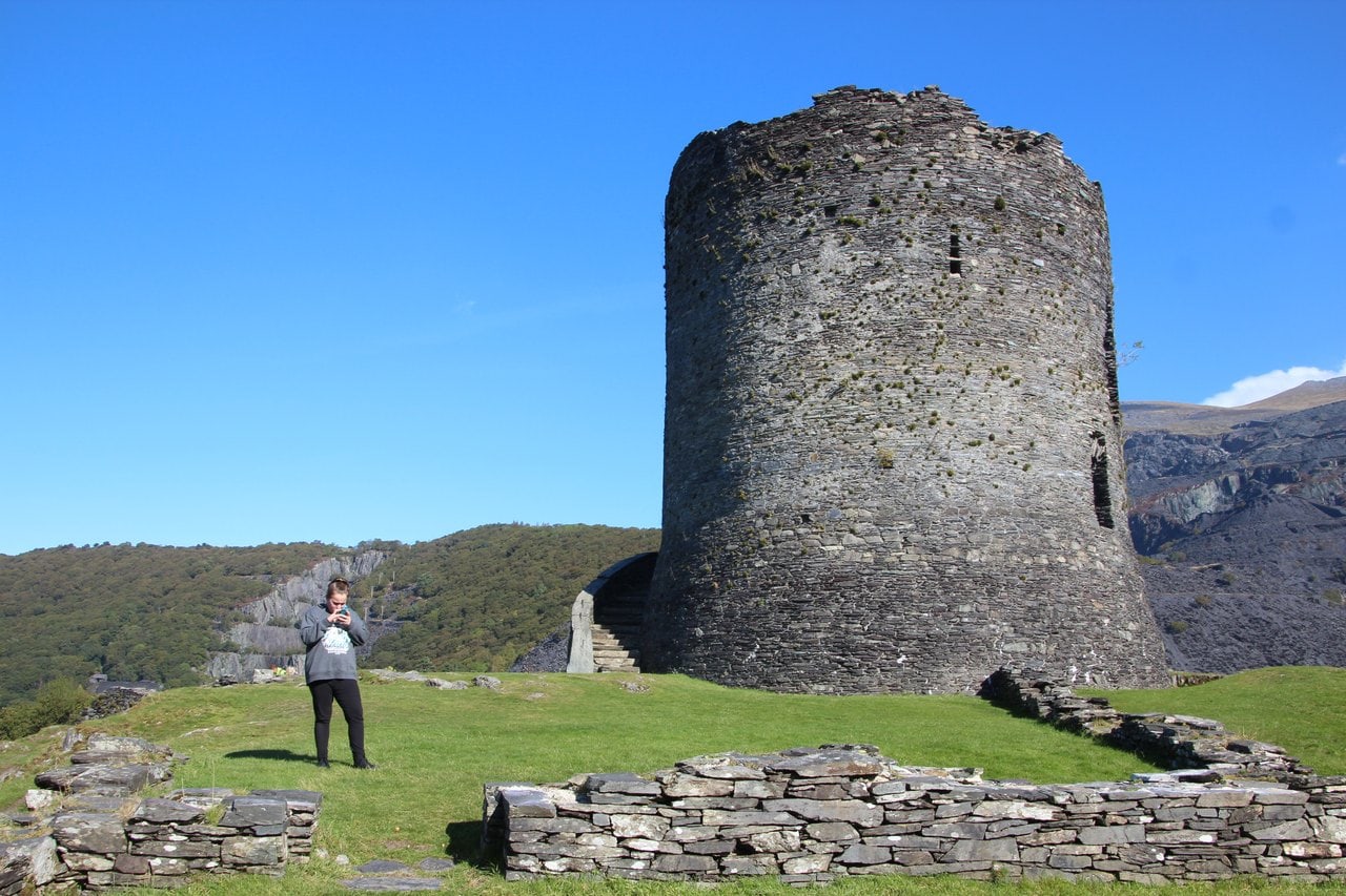 A tall round stone tower under blue sky with a flight of stone steps against its left side, a woman standing on the grass and low slate walls in the foreground A tall round stone tower under blue sky with a flight of stone steps against its left side, a woman standing on the grass and low slate walls in the foreground