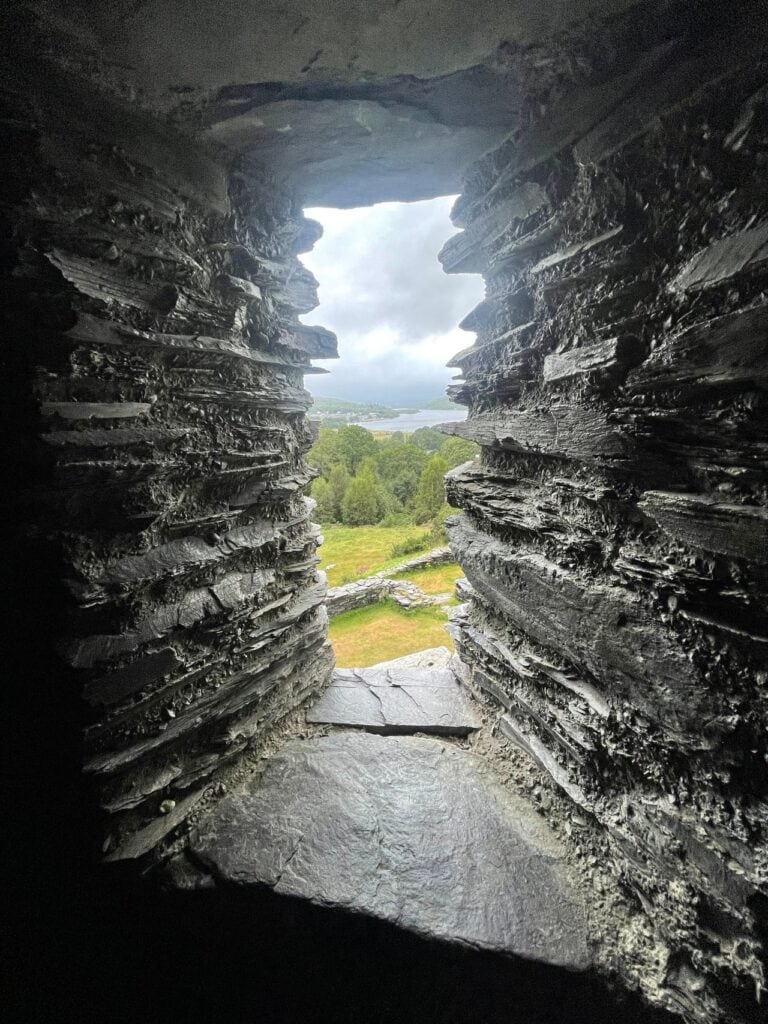 Dolbadarn Castle 10 A narrow deep window recess in a thick wall of thin, layered slate, opening onto green grass, trees and a glimpse of water