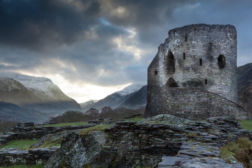 Dolbadarn Castle 13 A ruined round stone tower on the right above slate footings, under dark cloud, with snow-capped mountains and a misty valley to the left